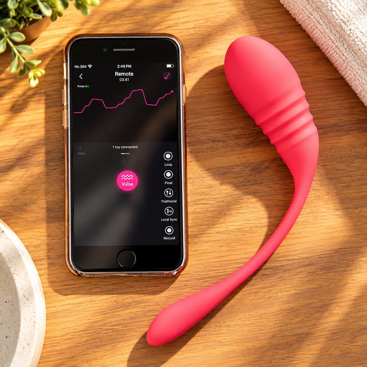 Pink rabbit vibrator on wooden surface next to an iPhone running a pulse-style app control screen